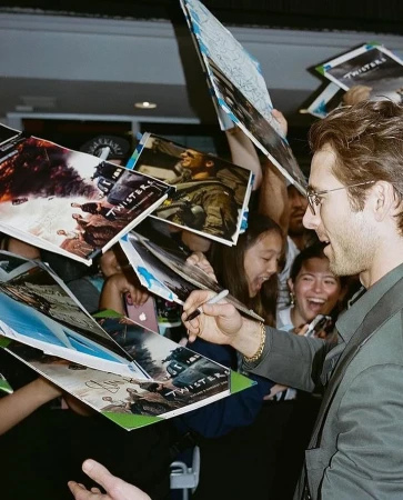 Actor Glen Powell signing autographs on 'Twisters' movie posters surrounded by fans at an event, smiling, wearing glasses.