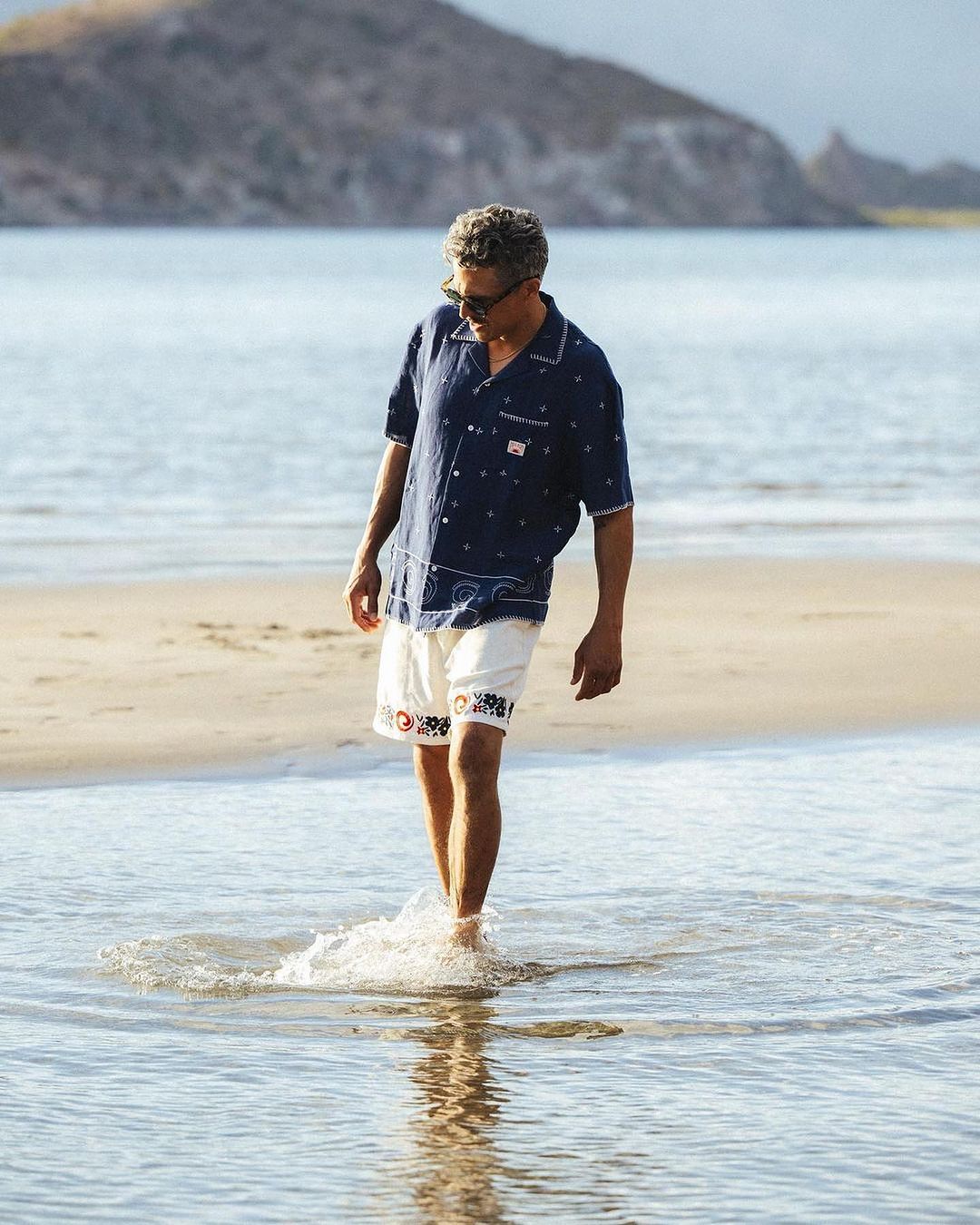 Man in casual summer outfit walking in the shallow ocean surf at the beach with mountains visible in the background on a sunny day.