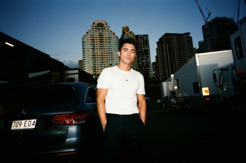 Portrait of a young man standing on a city street with buildings and commercial vehicles, wearing a white t-shirt and dark trousers.