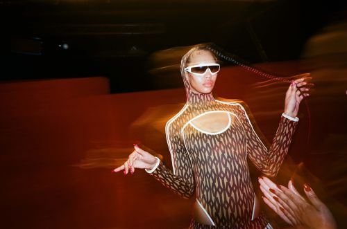 Adult woman with sunglasses and patterned bodysuit poses in an indoor setting with orange walls and another person's hands showing.