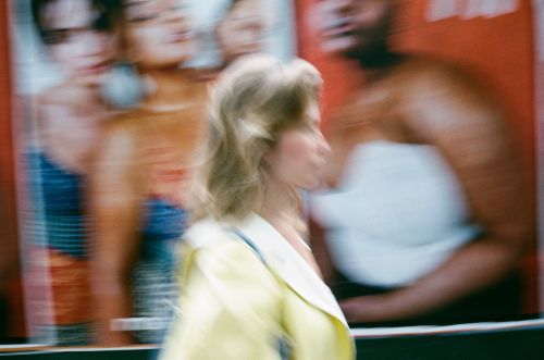 Blurred image of a woman walking in front of posters featuring blurred people, captured with a vintage photo effect and motion blur.