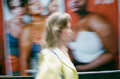 Blurred image of a woman walking in front of posters featuring blurred people, captured with a vintage photo effect and motion blur.