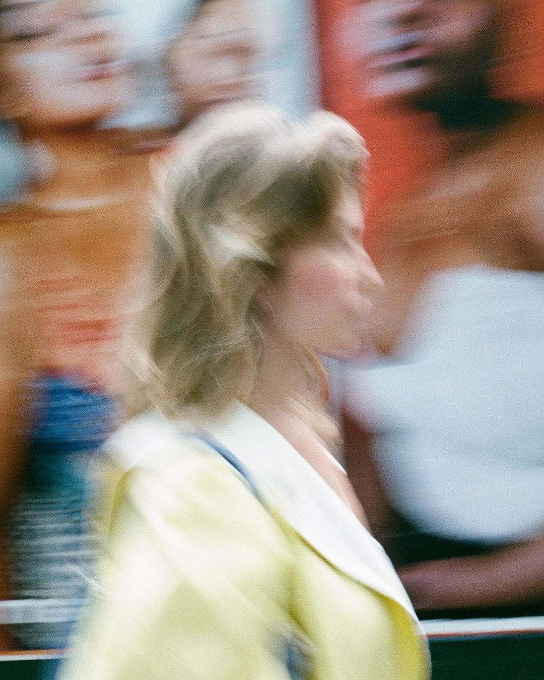 Motion-blurred shot of a woman with blond hair wearing a yellow jacket, walking past an out-of-focus advertisement on a city street.