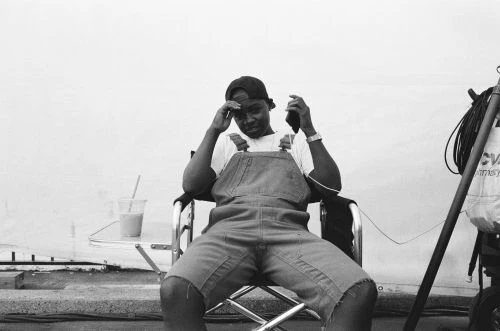 Young adult sits in director's chair wearing overalls, holding phone, with drink and equipment nearby, against a white backdrop.