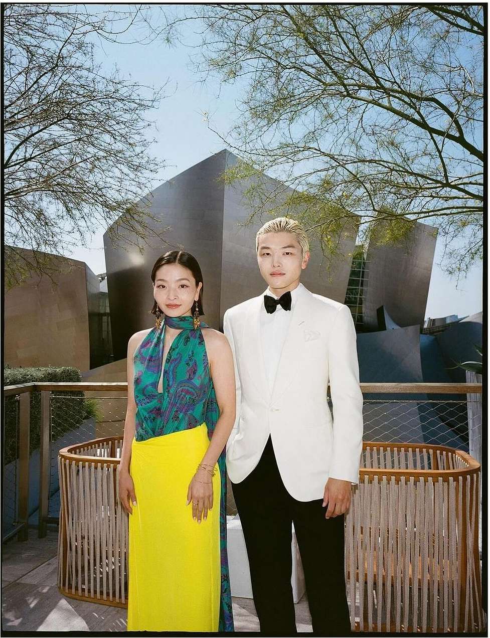 A formally-dressed couple poses outdoors in front of an architecturally modern building with bare trees against a blue sky backdrop.