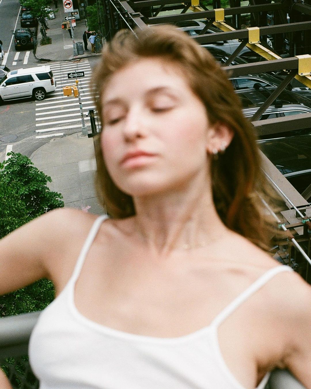 Outdoor portrait of a young adult female in a white tank top, eyes closed, posing on a bridge overlooking city traffic.