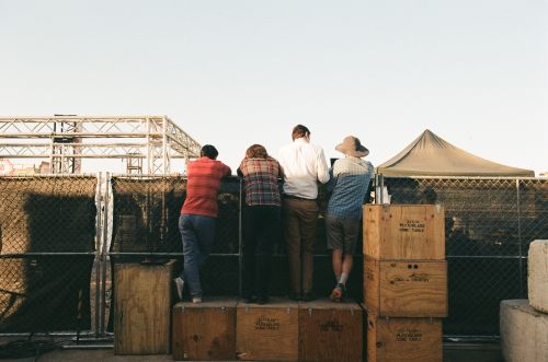 Four adults are standing on wooden crates looking over a chain link fence outdoors under a bright sky, observing an event space.