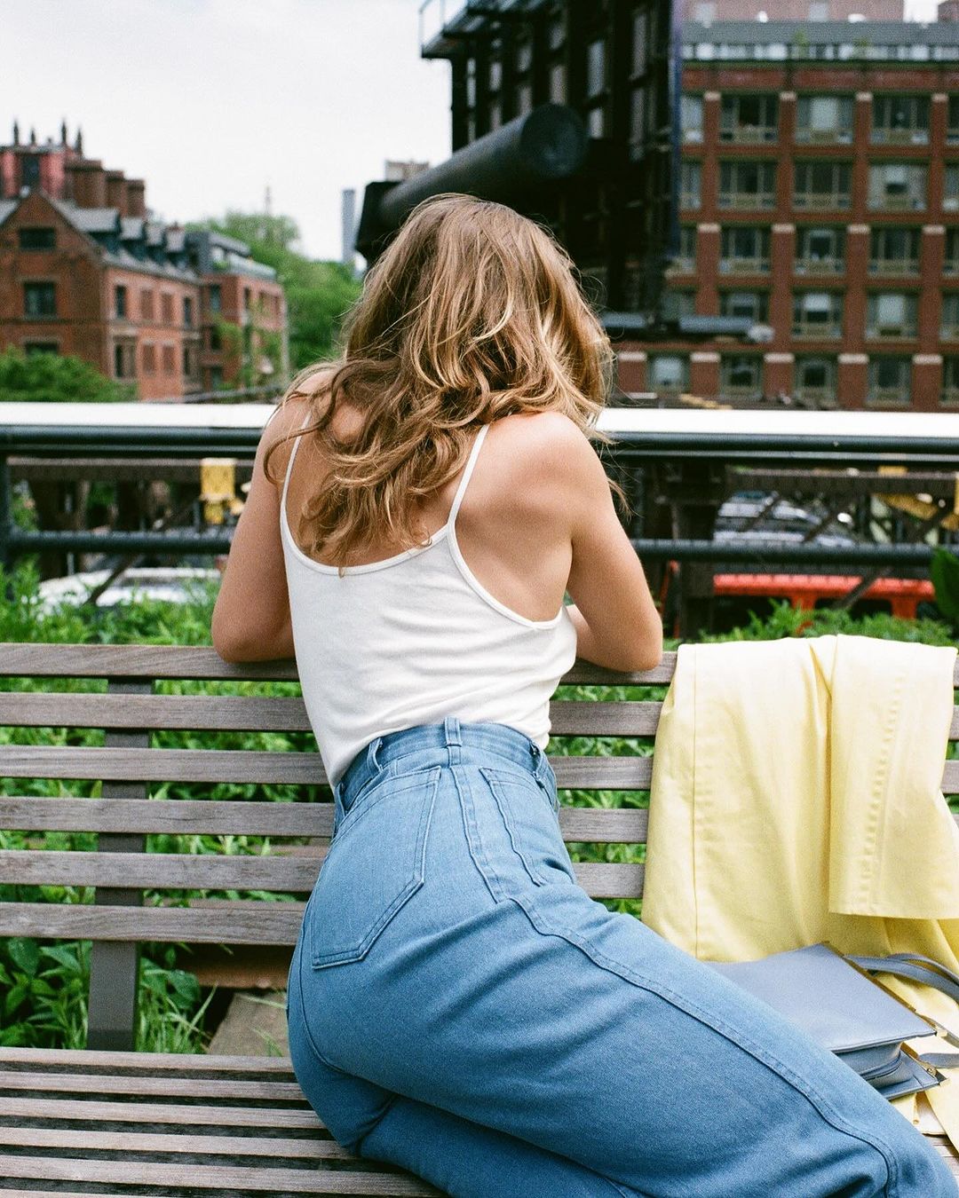 Blonde woman with light blue jeans sits on bench, back to camera. View of buildings, blue sky and foliage. Light yellow jacket nearby.