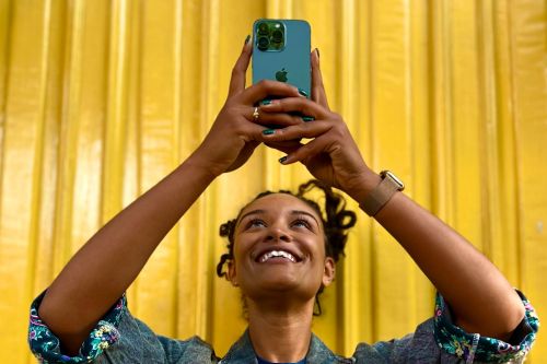 Smiling woman in a denim jacket holds up a smartphone against a yellow, vertically-paneled background, arms raised, looking up.