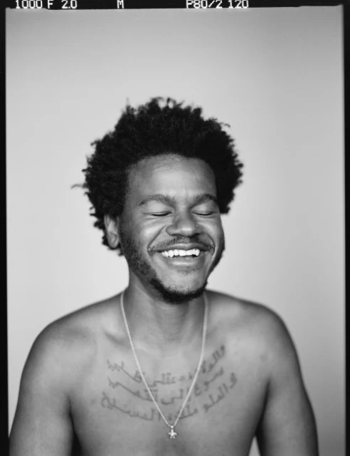 Close-up portrait of a smiling adult male with dark hair and a silver necklace, set against a simple grey backdrop in black and white.