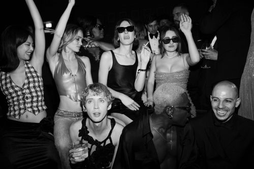Group of young adults gather at an indoor party in dark lighting, posing and smiling while wearing party outfits in a social setting.