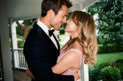 Close-up of a smiling couple in formal wear embracing outdoors. The woman wears a sleeveless pink dress and the man a black tuxedo.