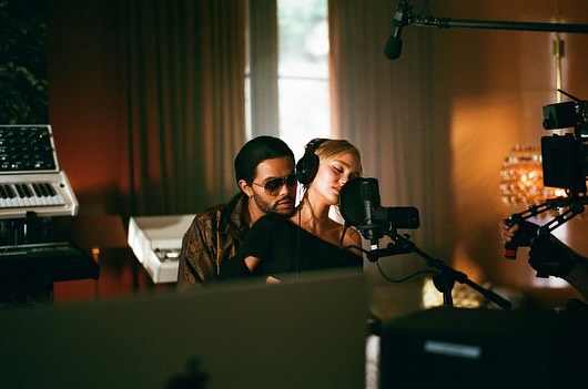 Male and female musicians collaborate in a recording studio, singing into a microphone with audio equipment and a camera visible.