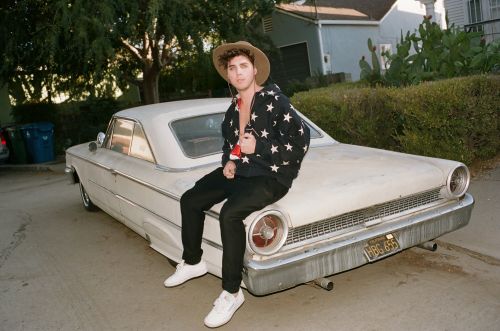 Young adult in casual wear sits on the hood of a vintage white car parked on a sunny residential street with green foliage.