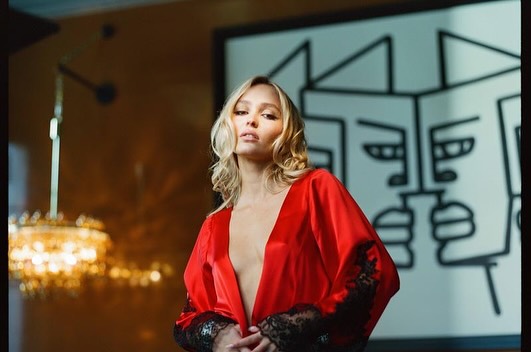 Adult female in a red robe poses indoors with blonde wavy hair, looking at the camera with art and a chandelier in the background.