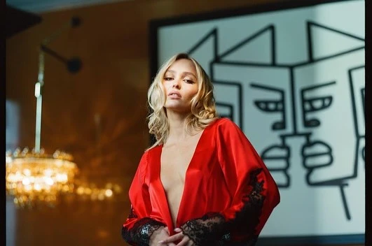Adult female in a red robe poses indoors with blonde wavy hair, looking at the camera with art and a chandelier in the background.