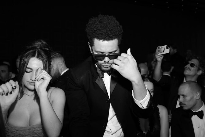 Black-and-white photo of a fashionable adult man in a suit and sunglasses, along with an adult woman, at a dimly lit social event.