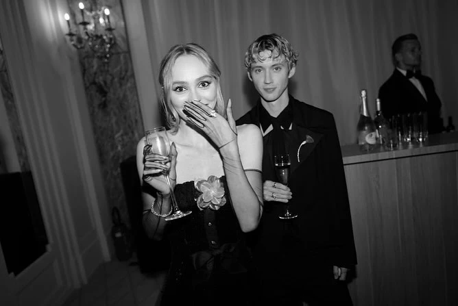 Two formally dressed adults, one holding a champagne flute, stand indoors near a curtained window, champagne bottles, and glasses.