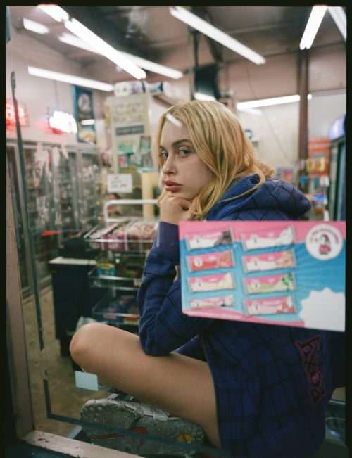Young woman poses in a store with snack cakes, wearing a hoodie and sneakers, captured in a casual, candid style with natural lighting.
