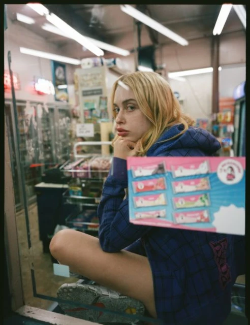 Young woman poses in a store with snack cakes, wearing a hoodie and sneakers, captured in a casual, candid style with natural lighting.