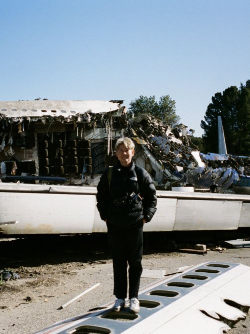 Adult stands in front of wreckage of airplane, wearing black jacket, pants, and shoes, with camera around neck, under clear sky.