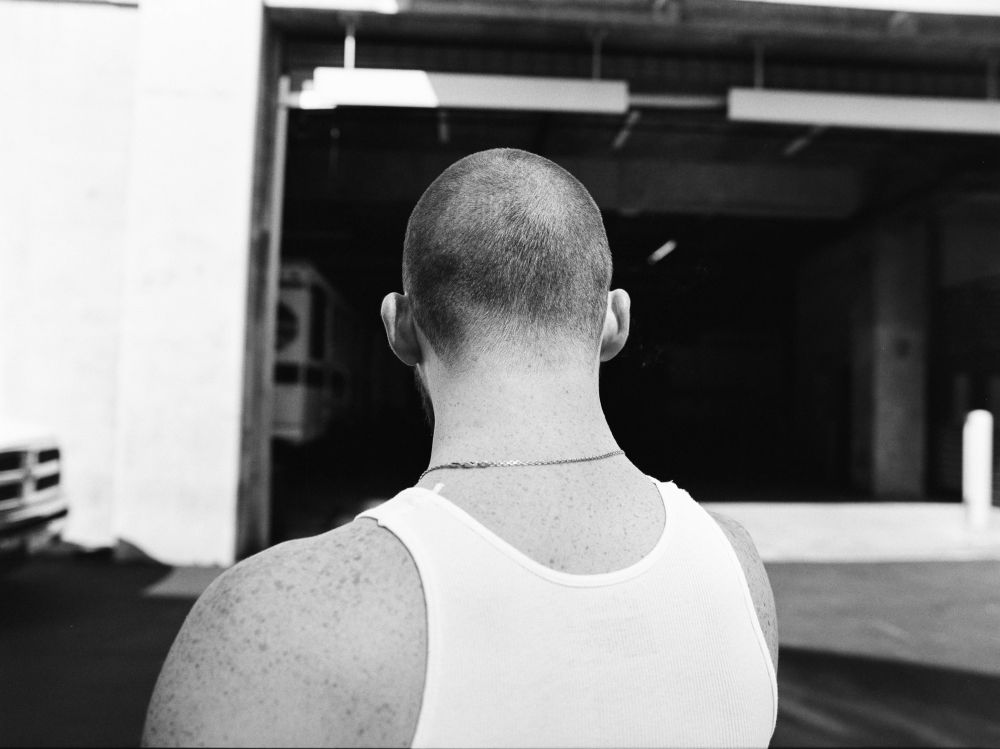 Back view of adult male with buzz cut, wearing a white tank top, standing in an urban setting, with a garage in the background.