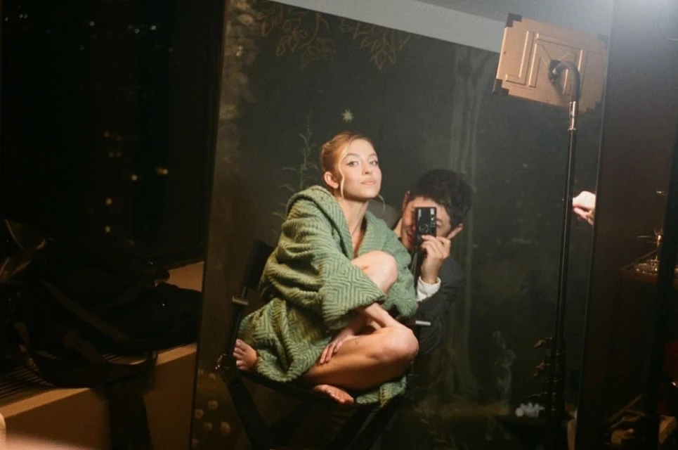 Blonde woman in a green robe poses in front of a mirror, reflecting a man holding a camera in an indoor setting.