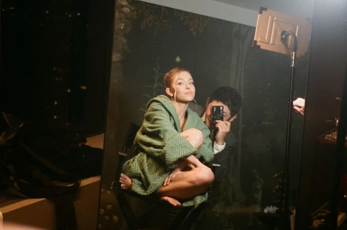 Blonde woman in a green robe poses in front of a mirror, reflecting a man holding a camera in an indoor setting.