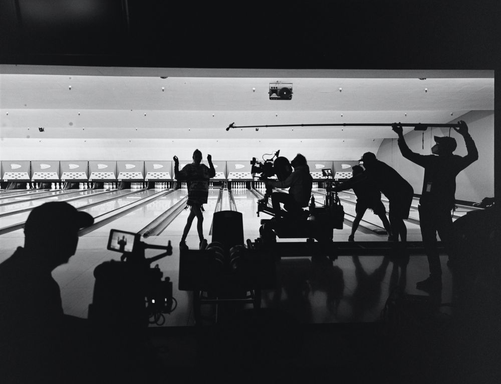 Filming a scene at a bowling alley with a camera crew, actors, and equipment visible in silhouette. Bright lighting and a long lane are present.