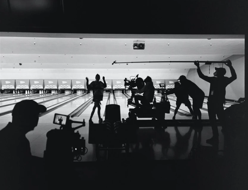 Filming a scene at a bowling alley with a camera crew, actors, and equipment visible in silhouette. Bright lighting and a long lane are present.