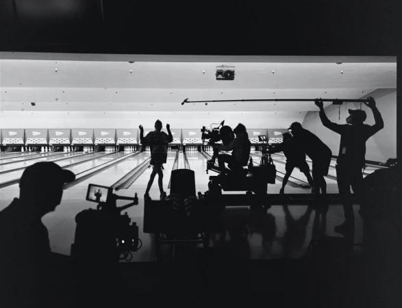 Filming a scene at a bowling alley with a camera crew, actors, and equipment visible in silhouette. Bright lighting and a long lane are present.