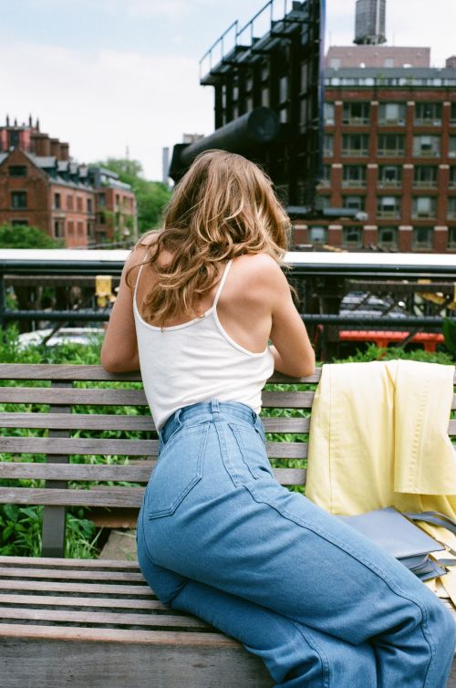 An adult woman in casual wear sits on a wooden bench in an urban park with buildings in the background on a sunny day.