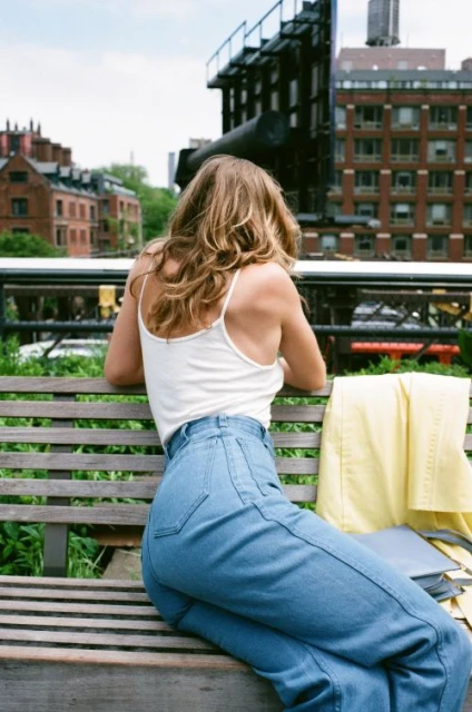 An adult woman in casual wear sits on a wooden bench in an urban park with buildings in the background on a sunny day.