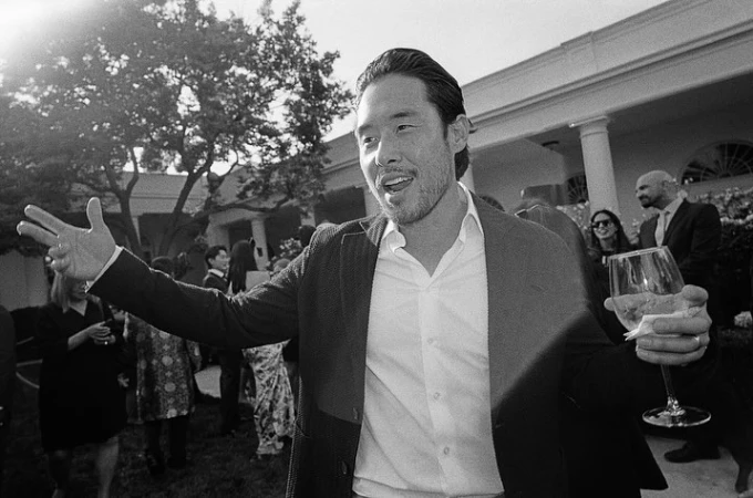 Black and white shot of an Asian man in a suit, smiling with his arm raised, holding a wine glass in front of a crowd outdoors.