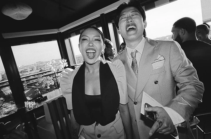 Two smiling adults, one man and one woman, indoors at a daytime event with a cityscape view, wearing a suit jacket and blouse. 