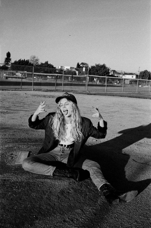 Black and white photo of a woman sitting on the ground with her arms raised, wearing jeans, a jacket, and a cap in an outdoor setting.