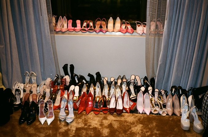 A large collection of high heels and shoes arranged on a floor and a windowsill between curtains.