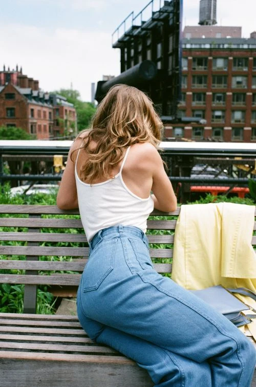 Woman with long blonde hair wearing a white tank top and blue jeans seated on a wooden park bench facing away.