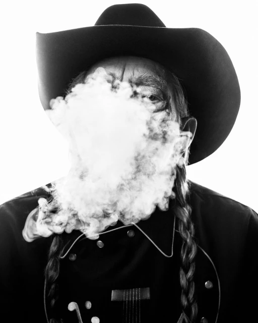 Black and white portrait of an adult male wearing a cowboy hat, partially obscured by smoke in front of a white backdrop.