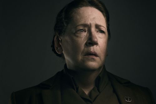 Close-up portrait of a mature woman with an emotional expression, wearing a dark green collared jacket with a silver pin, against a dark background.