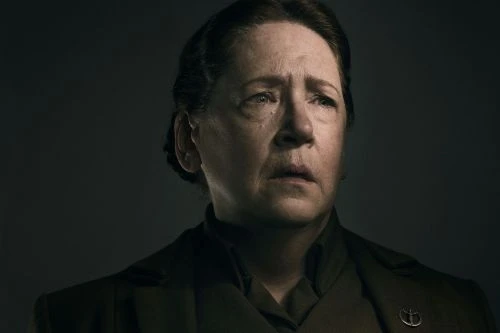 Close-up portrait of a mature woman with an emotional expression, wearing a dark green collared jacket with a silver pin, against a dark background.