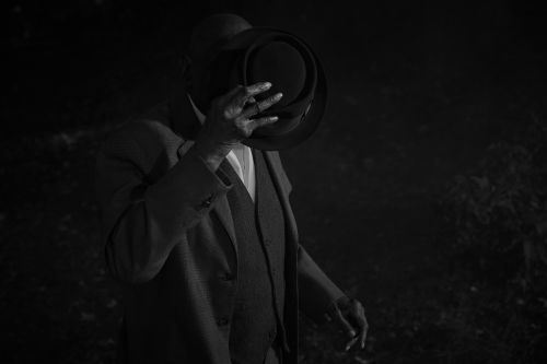 Monochrome portrait of an African-American man in a suit and hat, partially obscuring his face with the hat in a moody, dark setting.
