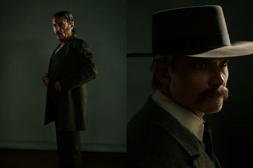 Dramatic side-lit portrait of two adult males wearing vintage suits and hats, one in a dark suit and the other in a hat.