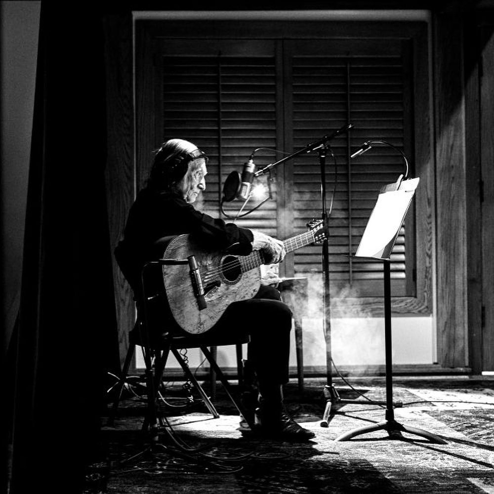 Black and white shot of an elderly man seated playing acoustic guitar in a music studio with a microphone and music stand.