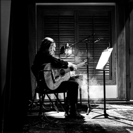 Black and white shot of an elderly man seated playing acoustic guitar in a music studio with a microphone and music stand.