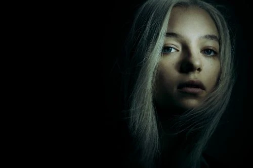 Close-up portrait of a young woman with long pale blonde hair and piercing blue eyes emerging from deep shadows.