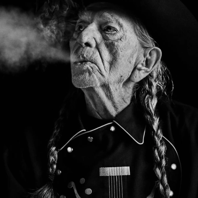 Monochrome portrait of an elderly man with braids wearing a Western-style shirt, a hat, and blowing smoke against a dark backdrop.