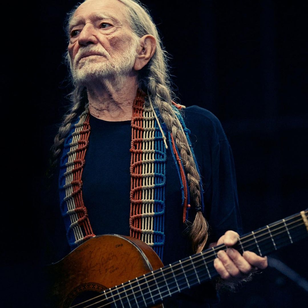 Willie Nelson with long braids holds his guitar in a medium shot against a dark background. He wears a colorful patterned strap.