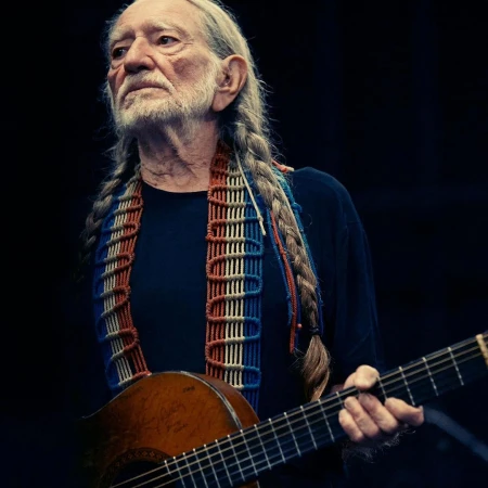 Willie Nelson with long braids holds his guitar in a medium shot against a dark background. He wears a colorful patterned strap.