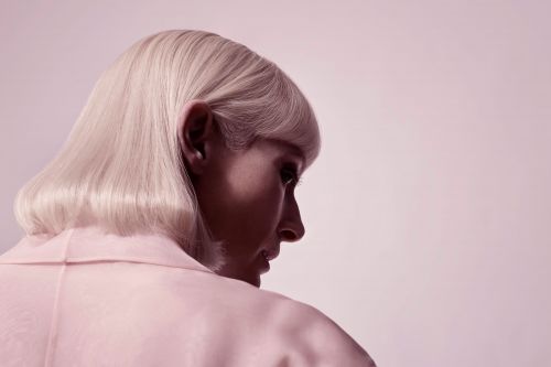 Head and shoulder shot of a woman with platinum blonde hair wearing a pale pink jacket, looking down in a photo studio.